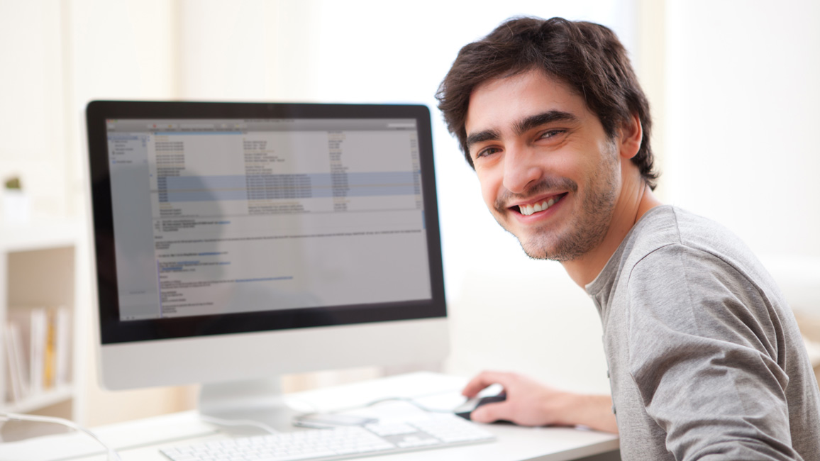 Ein junger Mann, sitzt an einem Schreibtisch vor einem PC, auf dessen Monitor eine Tabelle zu sehen ist, und dreht sich lächelnd zur Kamera.