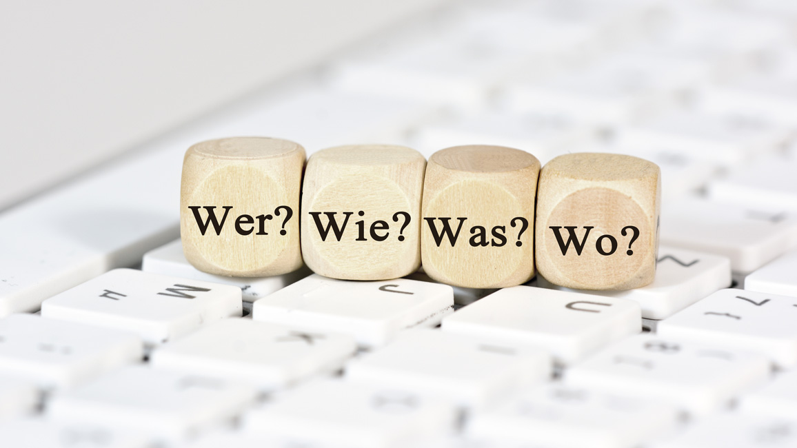 Four wooden dice showing the German words for „who“, „how“, „what” and “where”, placed on a computer keyboard.